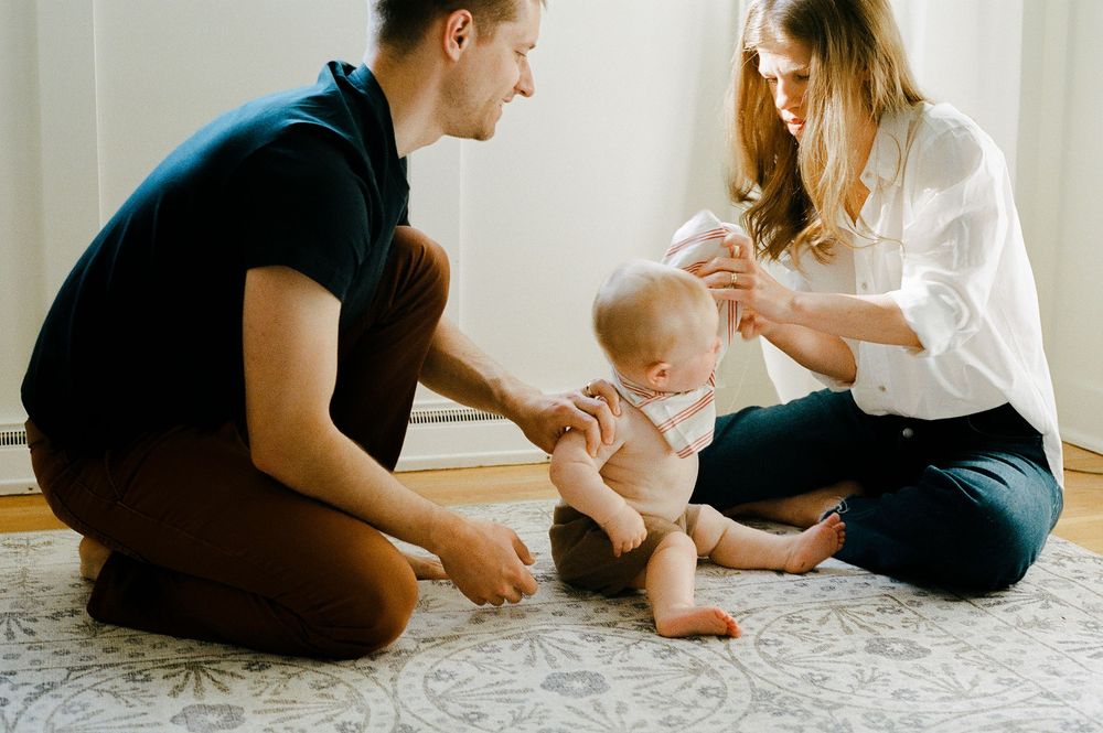 Parents sitting on floor with baby learning to stand while supporting each other on a patterned carpet.
