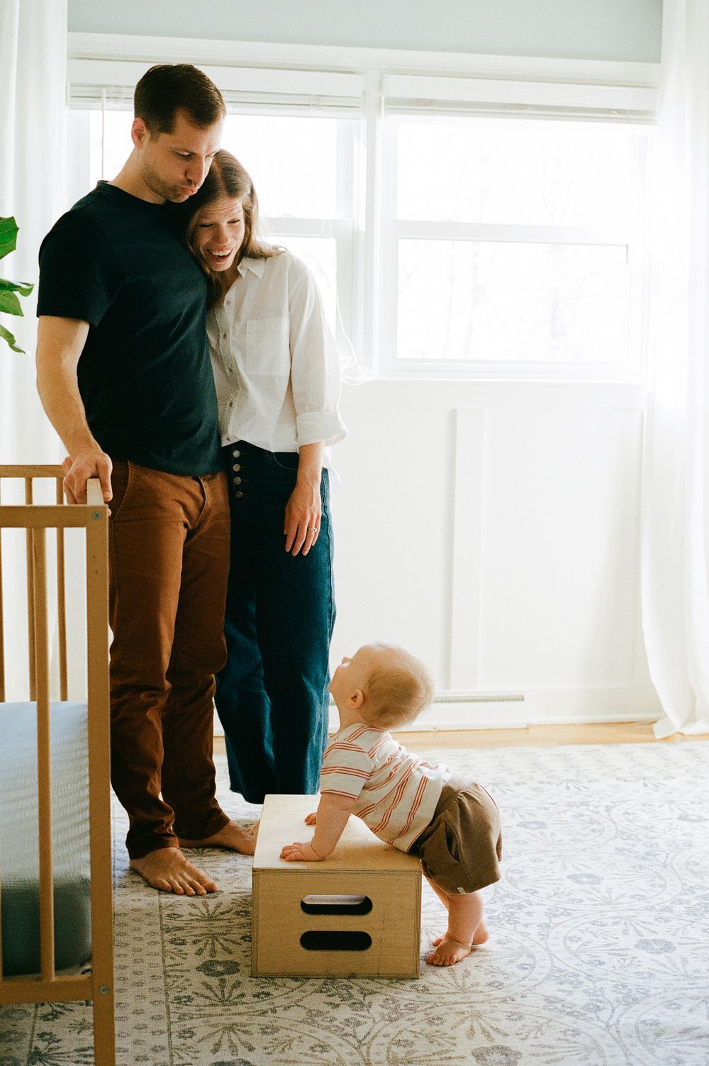 Toddler standing on wooden box while parents watch in bright room.