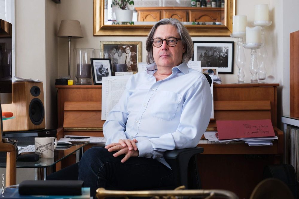 A person in glasses and a light blue button-down shirt sits in a home office setting with wooden furniture and framed artwork.
