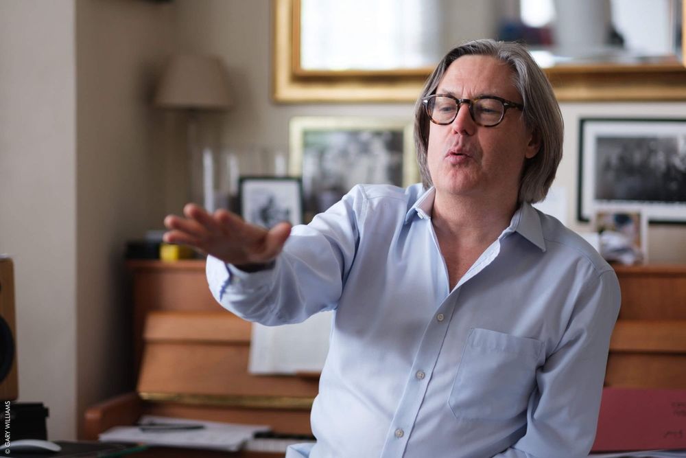 Person wearing a white collared shirt gestures with one hand while sitting in a room with framed photos on the wall.