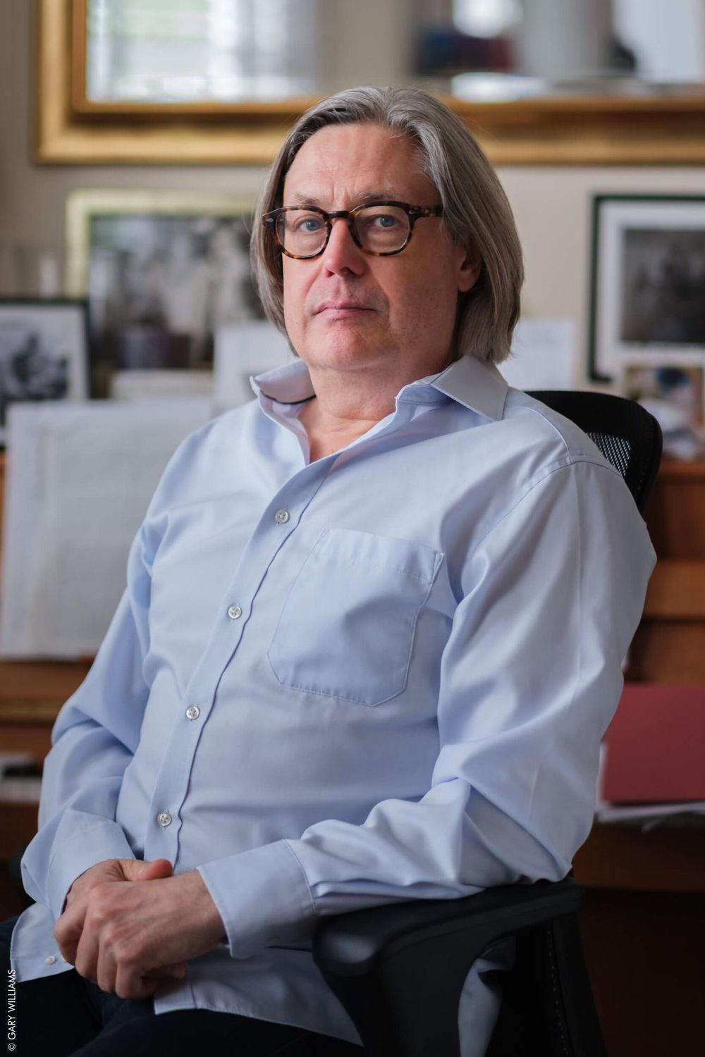A person wearing glasses and a light blue button-down shirt sits in an office with framed photos in the background.