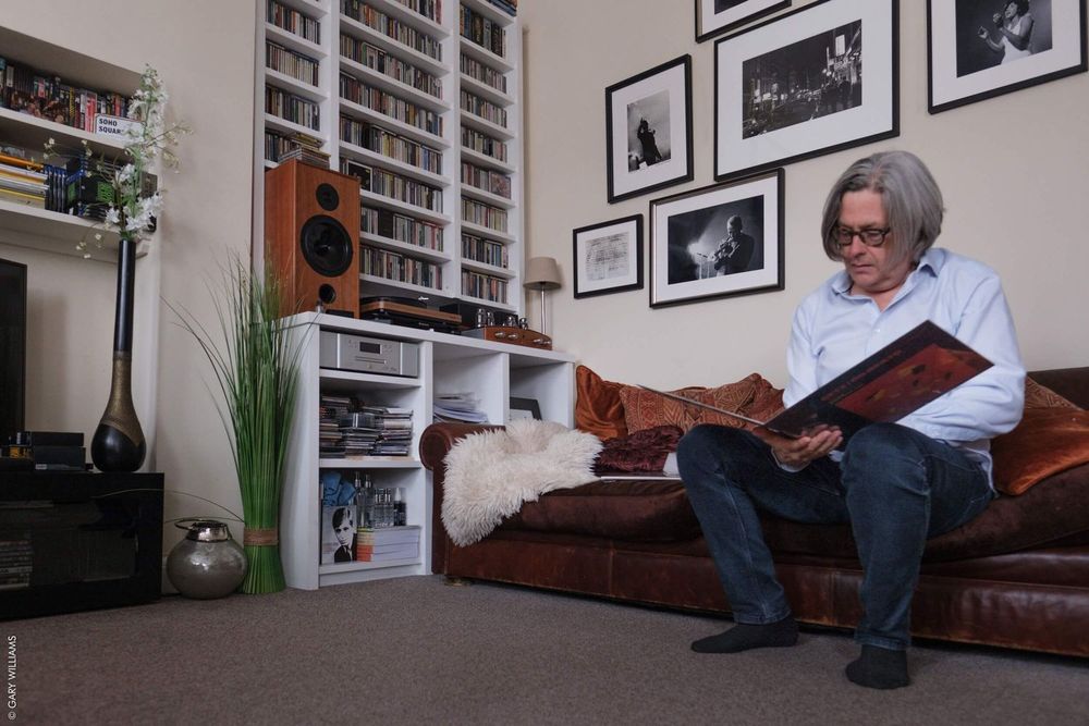 Someone reads a book while relaxing on a leather couch in a room with bookshelves and framed photos on the wall.