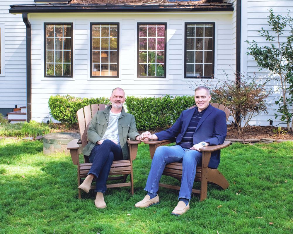 Two people sit relaxed in wooden Adirondack chairs on a green lawn in front of a white house with large windows.