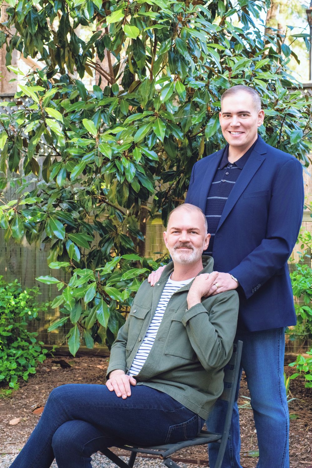 Two people pose in an outdoor garden setting with greenery and foliage in the background.
