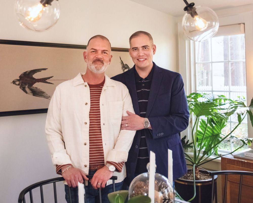 Two people stand together in a dining room with pendant lights, dark artwork and houseplants in the background.