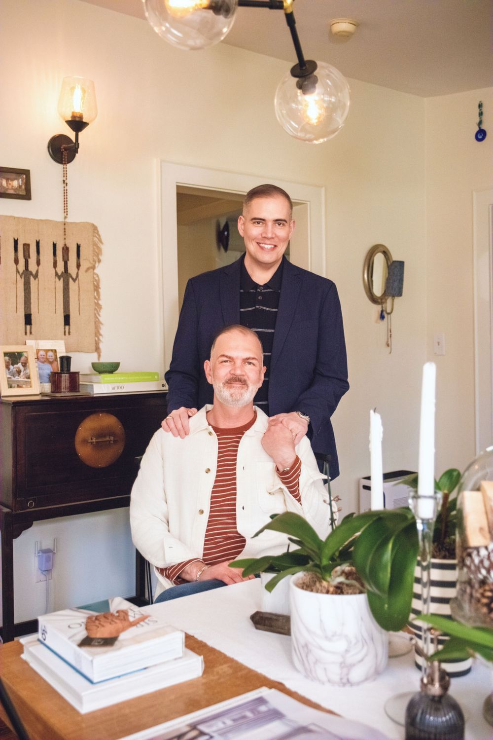 Two people sitting at a dining table with modern lighting fixtures, decor, potted plants and candles in a cozy interior setting.