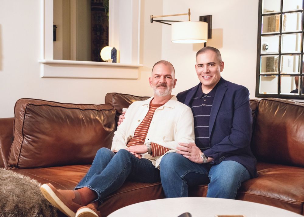 Two people sit together smiling on a brown leather couch in a bright living room with white walls.