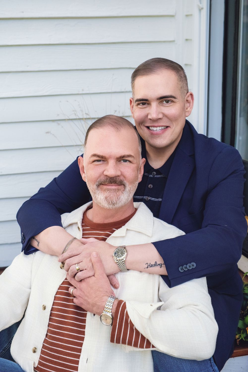 Two people embrace while posing for a portrait, one wearing a navy blazer and the other in a cream colored jacket.