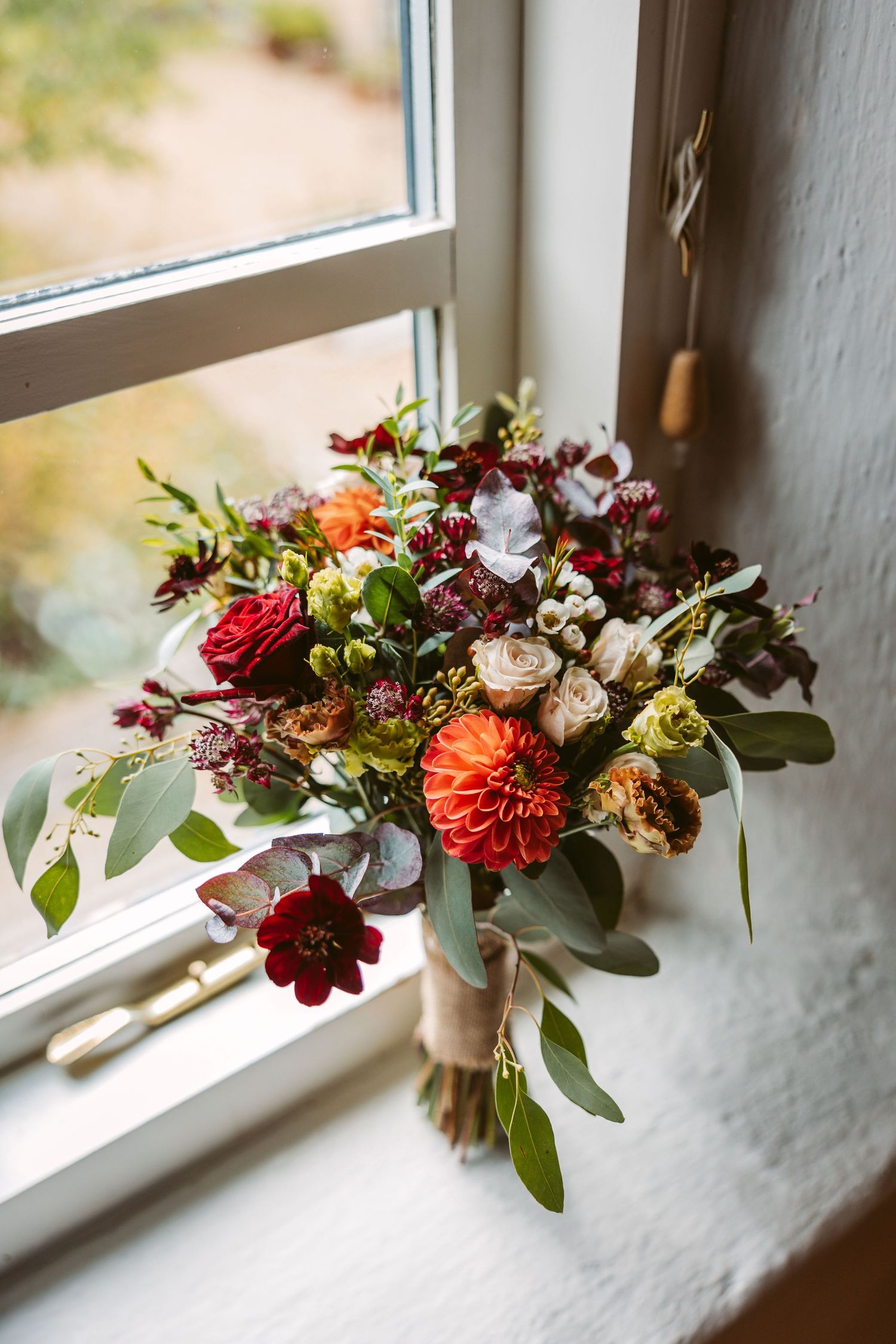 A vibrant autumn wedding bouquet with red dahlias, orange zinnias and eucalyptus sits on a white windowsill in natural light.