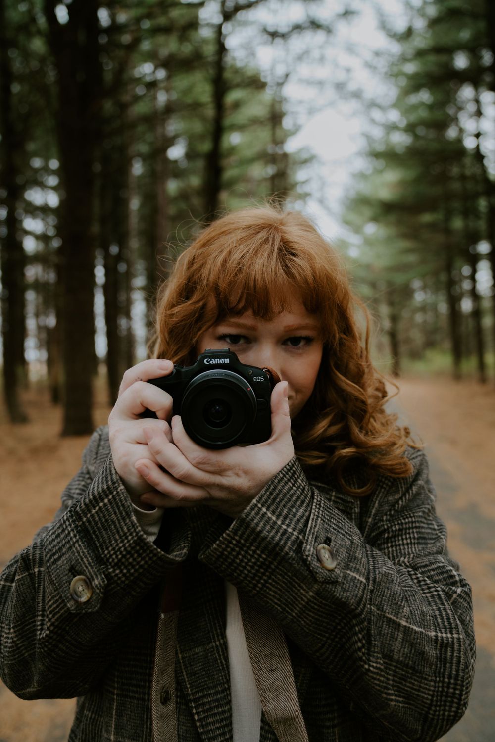 Photographer holding a Canon camera in a forest setting wearing a plaid coat.