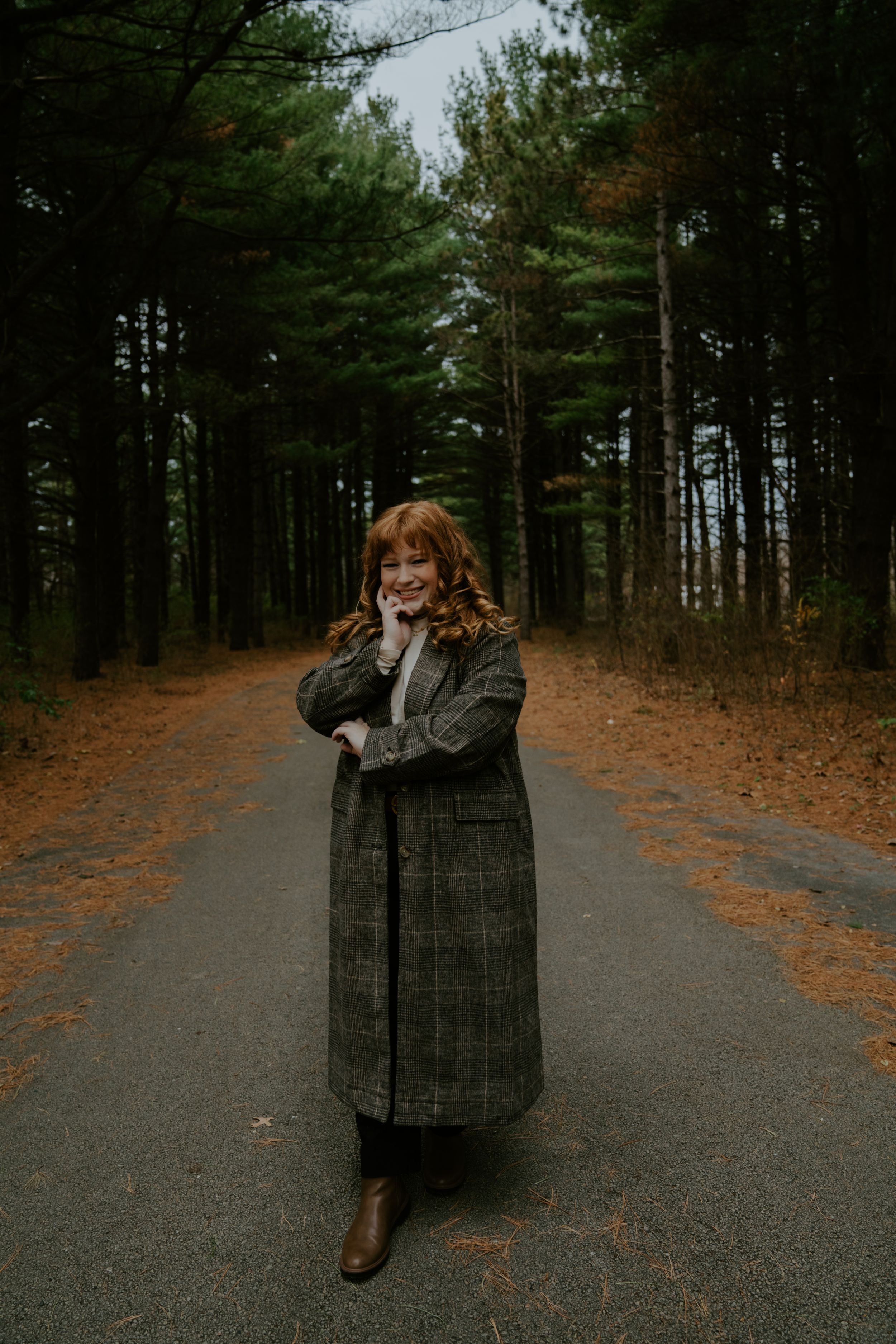 Figure in a checkered coat poses on a forest path surrounded by tall pine trees.