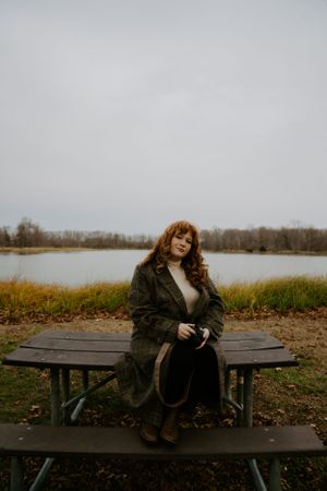 Person in plaid coat relaxing on wooden picnic table by autumn lakeside.