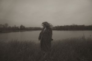 Moody atmospheric portrait of person in dark coat standing by lake in misty weather.