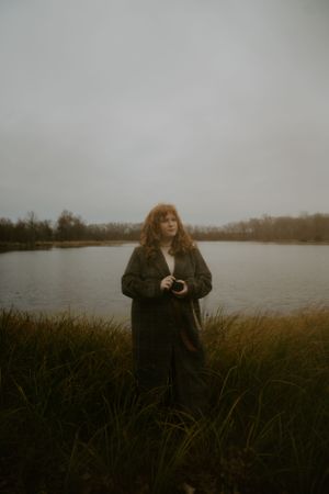 Person in dark winter coat holding camera by lakeside with tall grass in foreground.