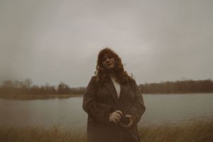 Figure with camera stands among tall grass by lake on overcast day.