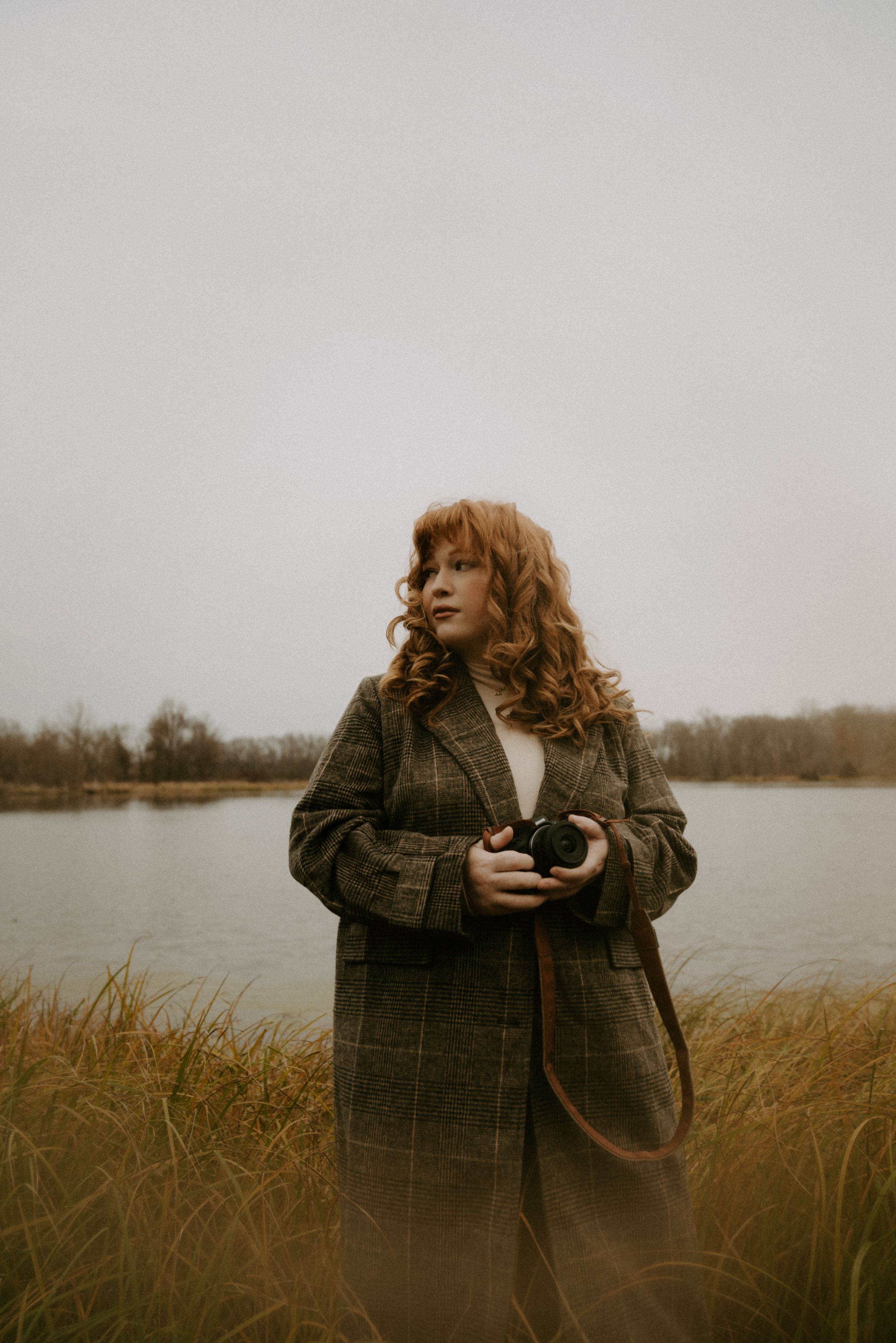Person in plaid coat holds vintage camera while standing in autumn field by water.