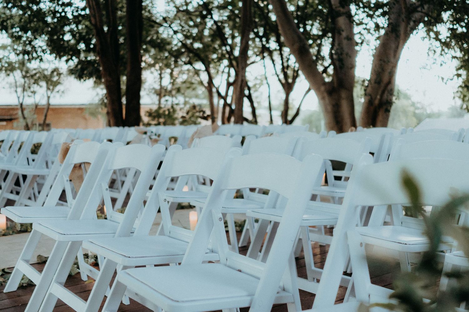 White wooden folding chairs arranged in rows outdoors for a ceremony beneath shaded trees.