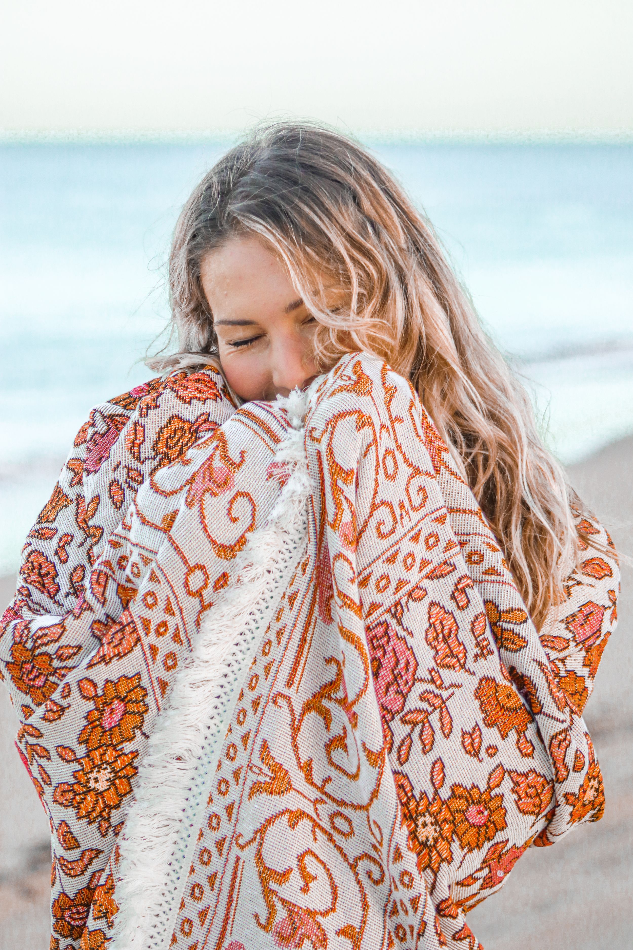 A person wraps themselves in a beautiful orange and white floral beach blanket by the ocean.