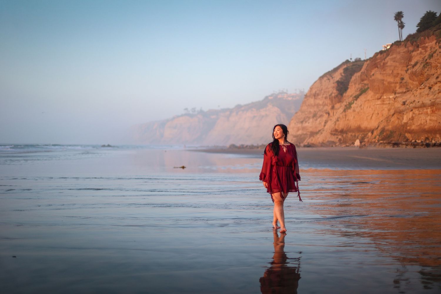 Woman in red dress walking on a misty beach at golden hour with cliffs in background.