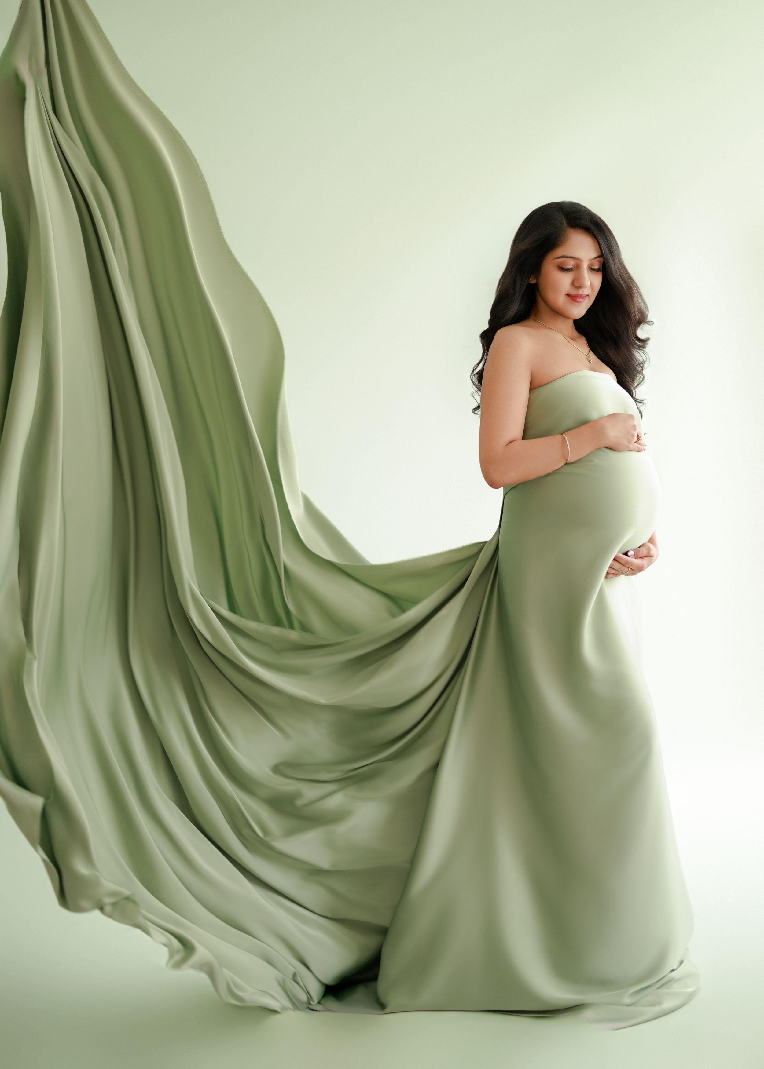 Elegant maternity portrait in flowing sage green dress against white background with billowing fabric creating dramatic effect.