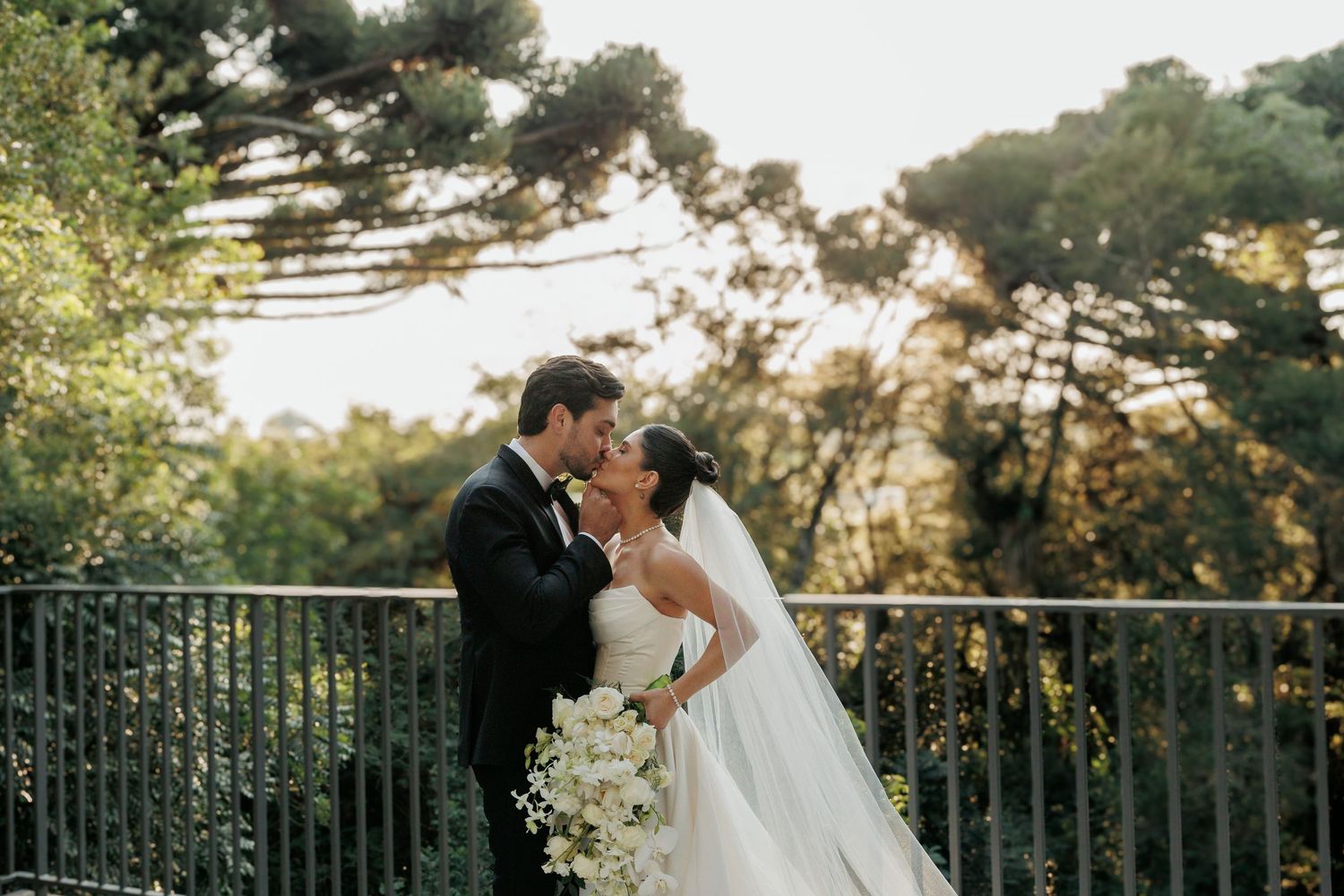 A romantic wedding ceremony moment captured outdoors at sunset with lush greenery and a metal railing backdrop.