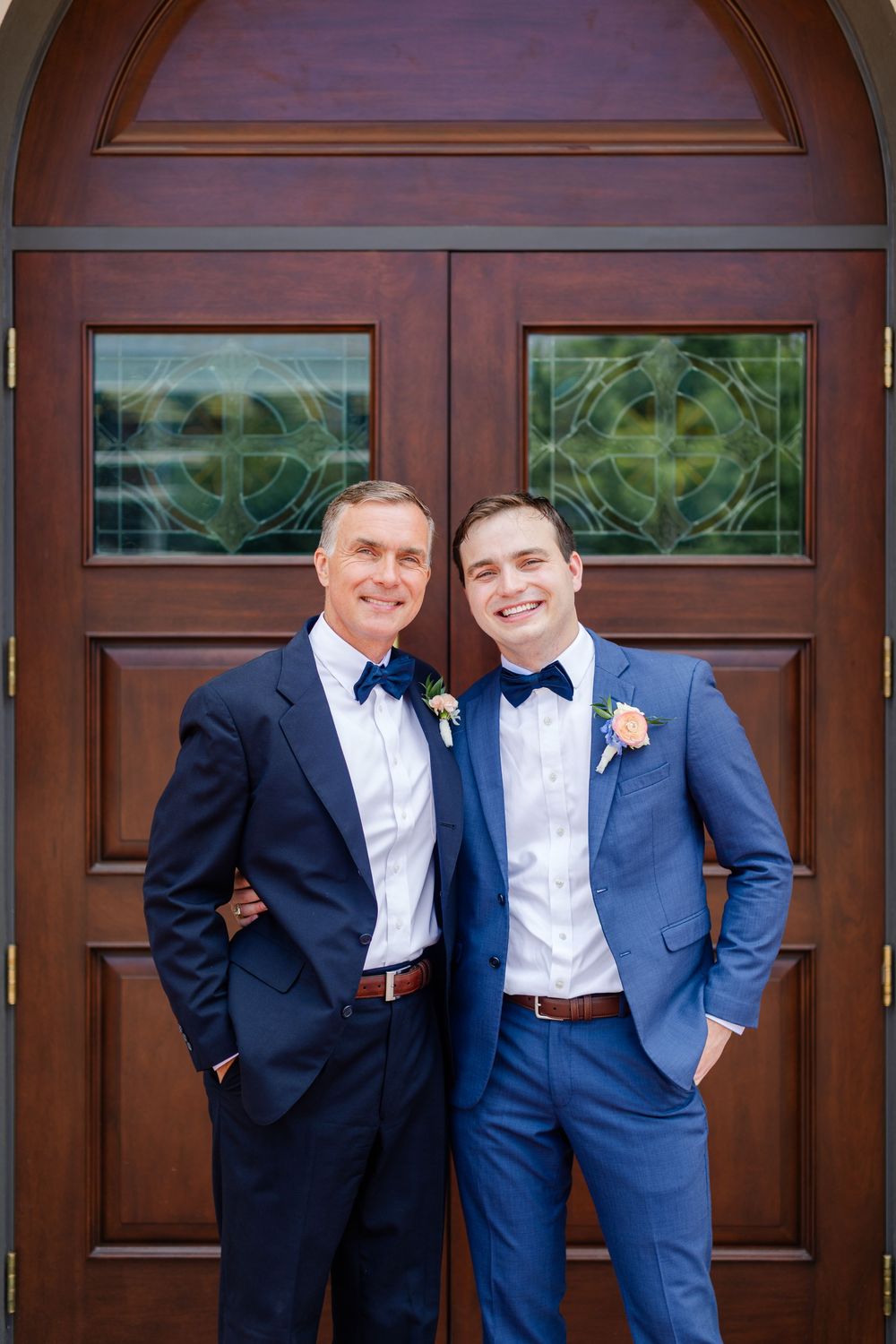 Two men in navy blue suits and bow ties pose in front of a wooden door with decorative glass panels.