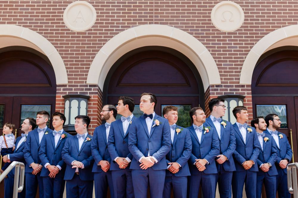 Groomsmen in matching blue suits lined up along a brick wall for formal event photos.
