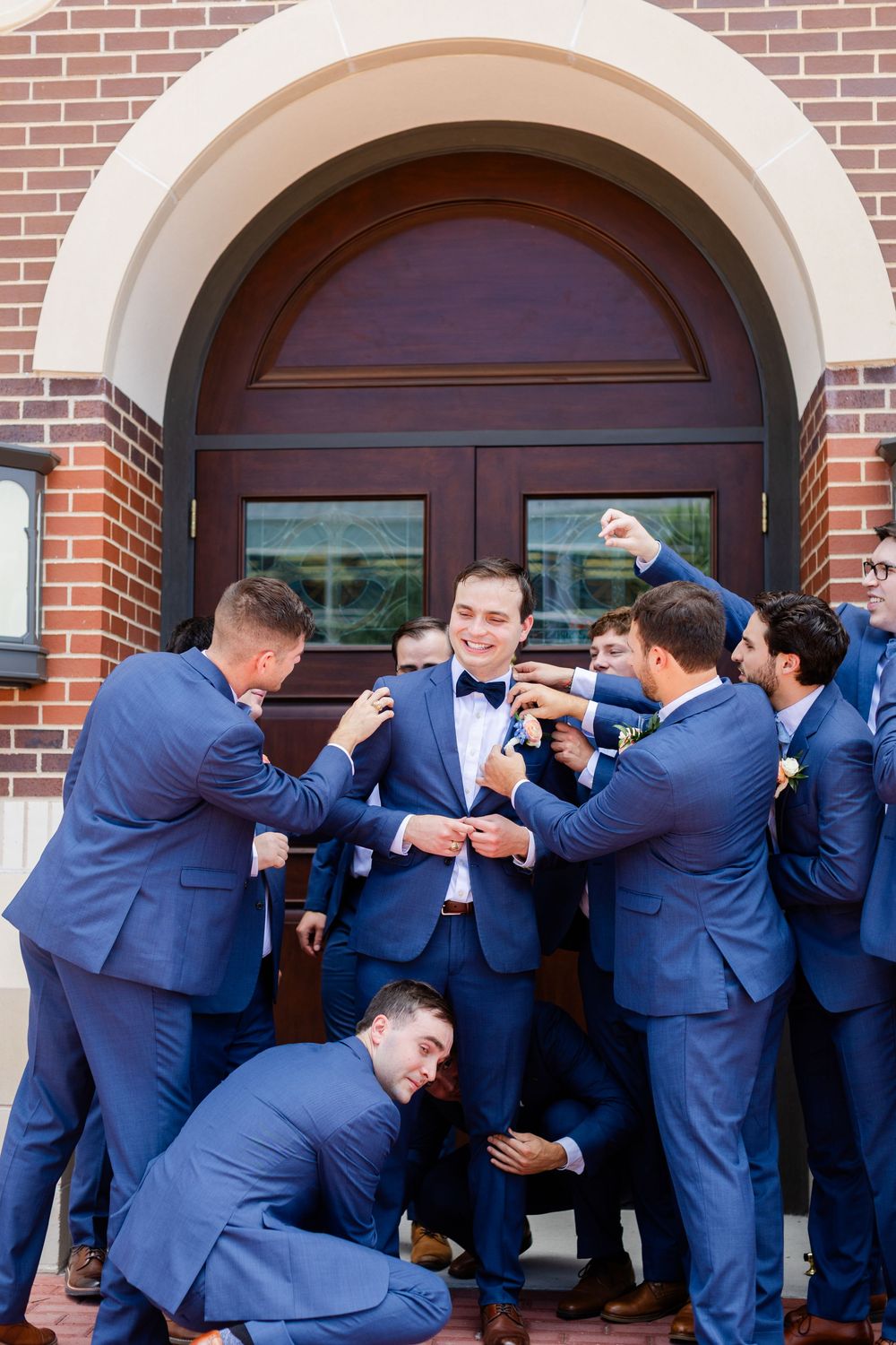 Groomsmen in navy blue suits playfully pose and celebrate together in front of a brick church entrance.