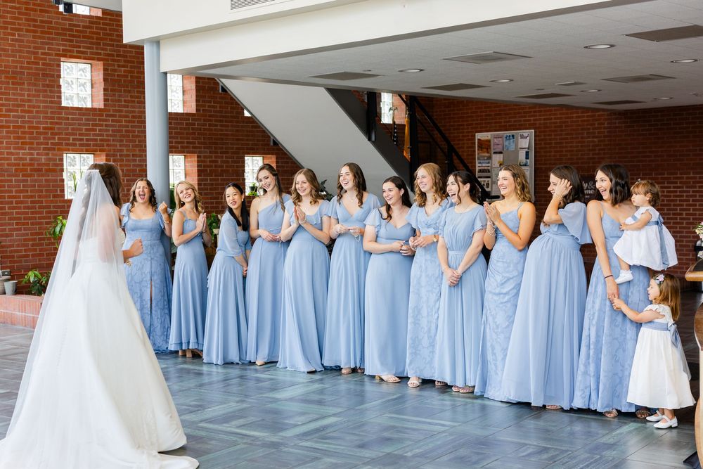 A group in floor-length light blue bridesmaid dresses stands in line indoors admiring a bride in a white gown.