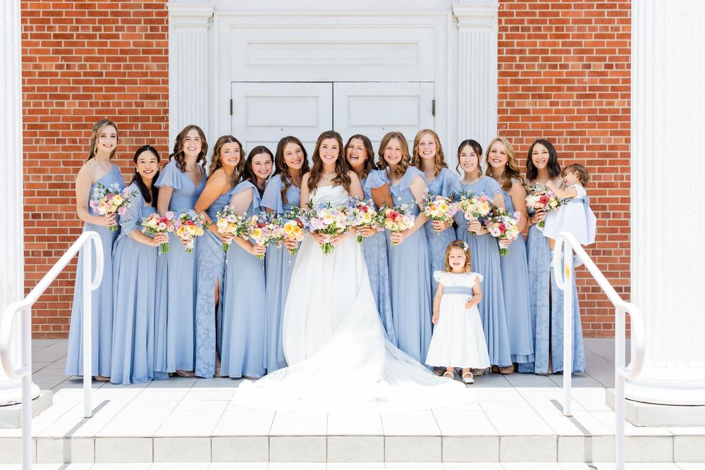 Wedding party in matching light blue bridesmaid dresses and white bridal gown pose on church steps with floral bouquets.