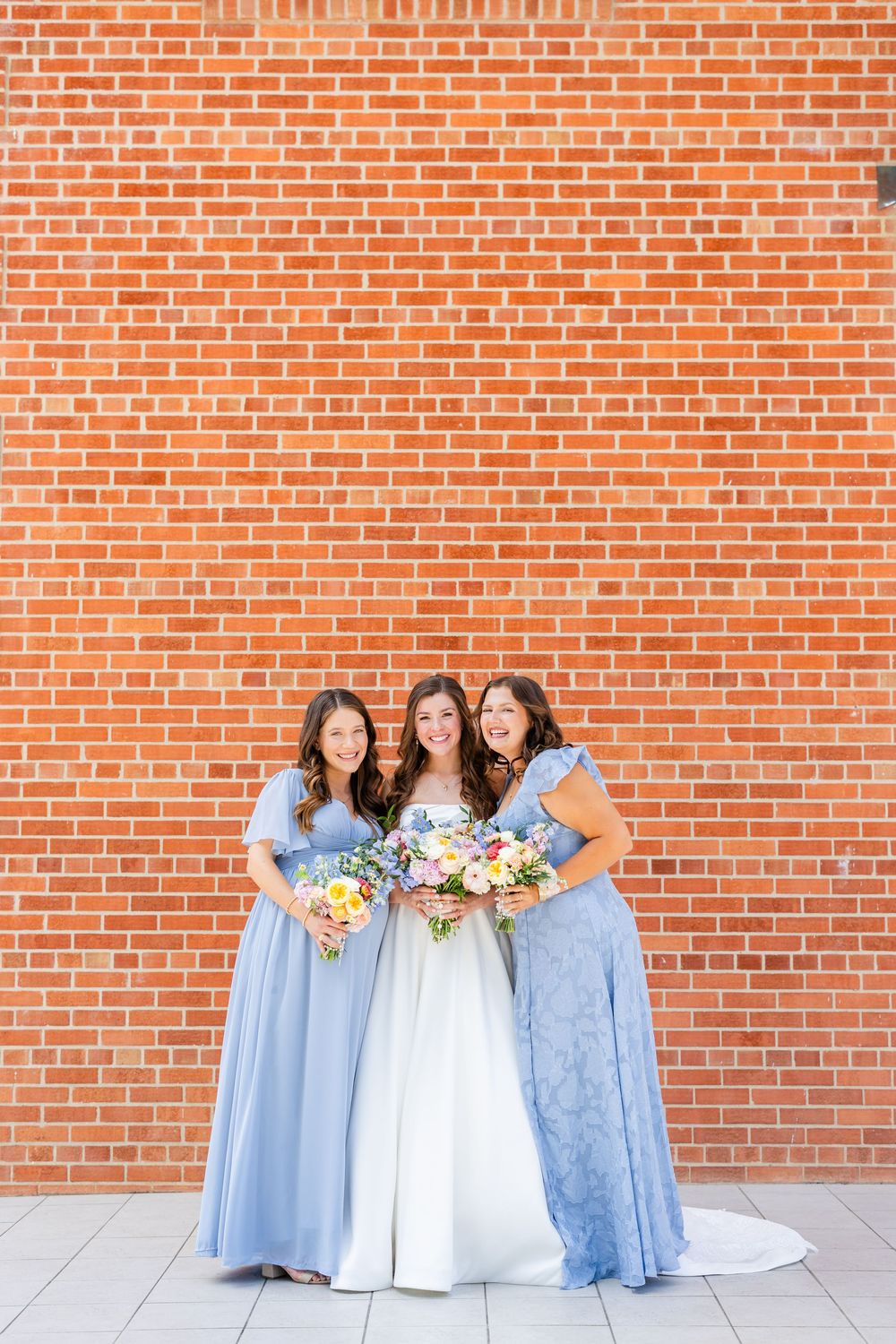 Bridal party in light blue bridesmaid dresses and white wedding gown pose against rustic brick wall with floral bouquets.
