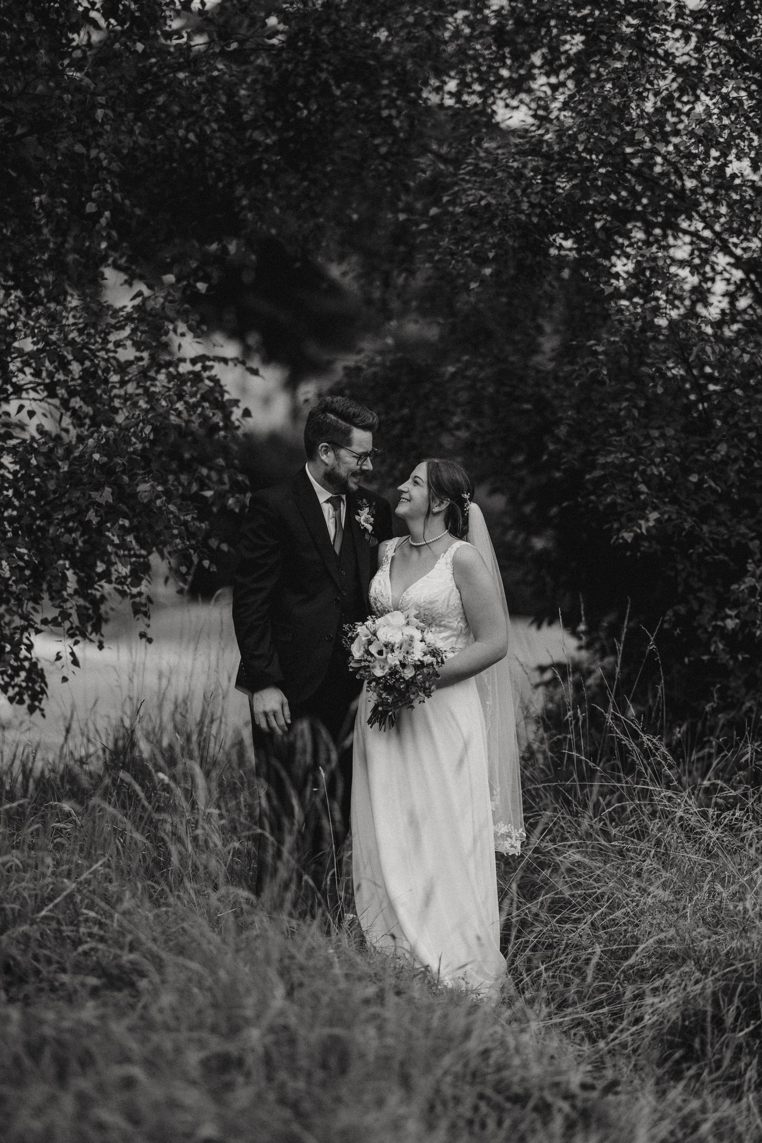 A romantic black and white wedding photo of a couple standing together in long grass surrounded by trees.