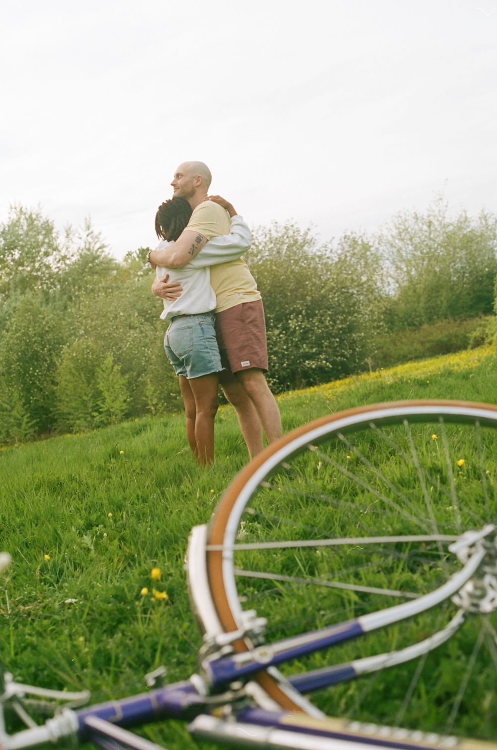 A bicycle lies in foreground while two people embrace in a sunny meadow.