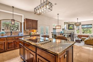 Elegant kitchen with granite island, rich wood cabinets, chandelier lighting, and open living area with large windows.