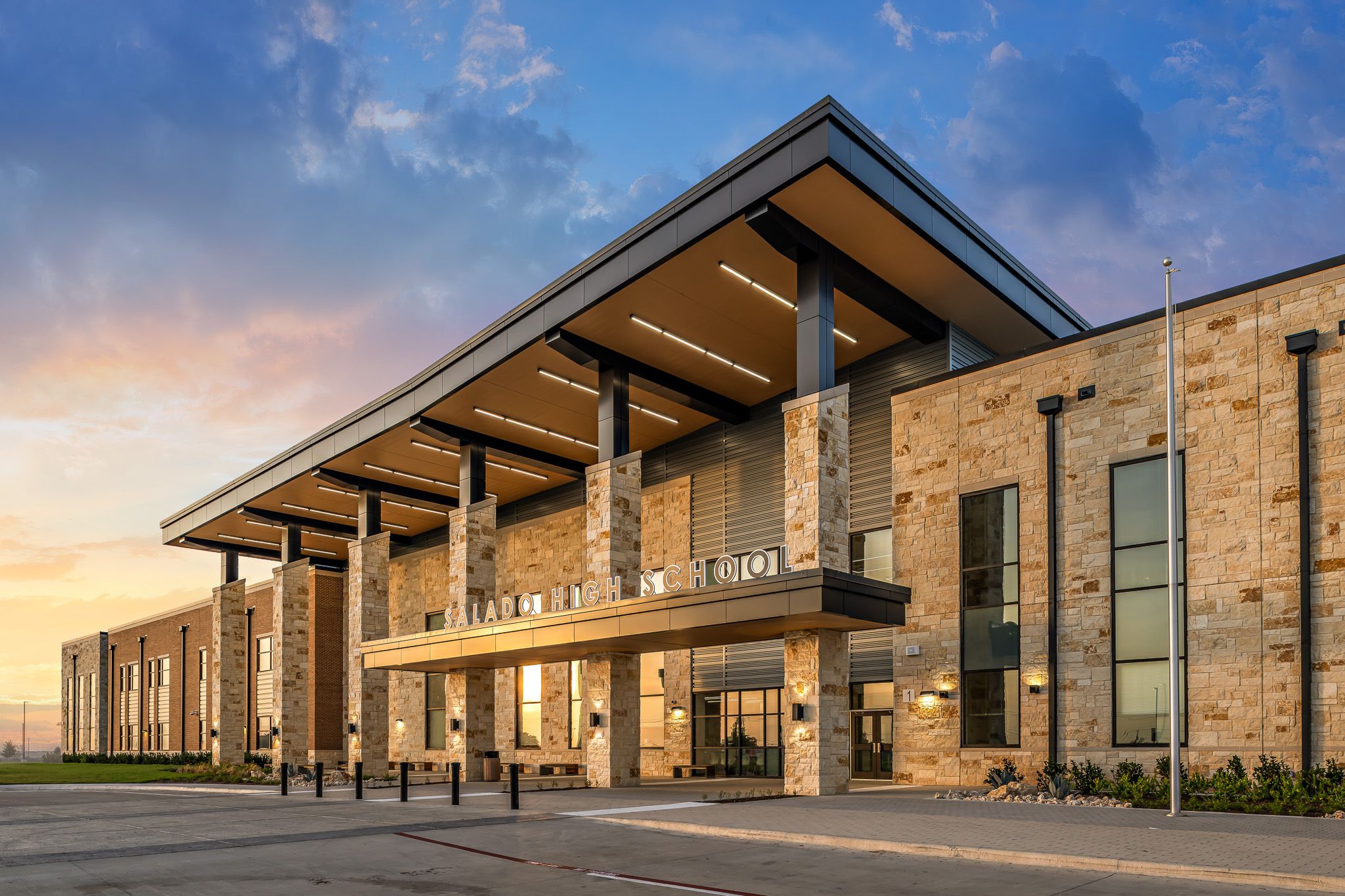 Modern stone building with large covered entrance, wood-accented roof, and tall pillars at sunset.