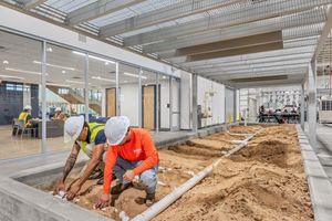 Two construction workers in hard hats lay pipes in a sand trench inside a modern building under construction.