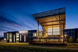 Modern corporate building exterior illuminated at dusk with glass facade and steel canopy against a blue twilight sky.