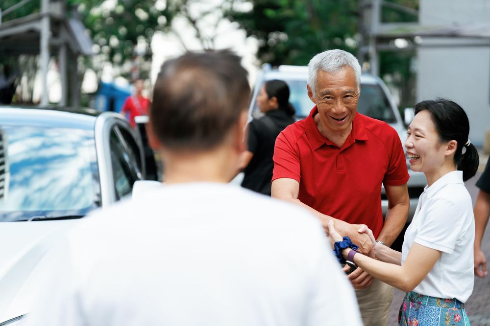 People in casual attire having a friendly conversation on a sunny street with cars parked nearby.