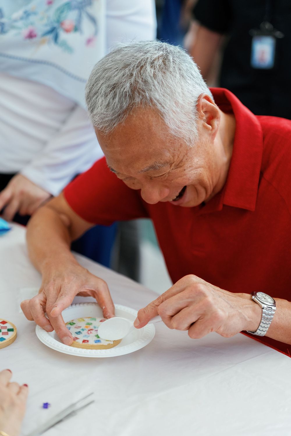 Person wearing a red polo shirt decorates a cookie with frosting at a white table, smiling while focused on their work.