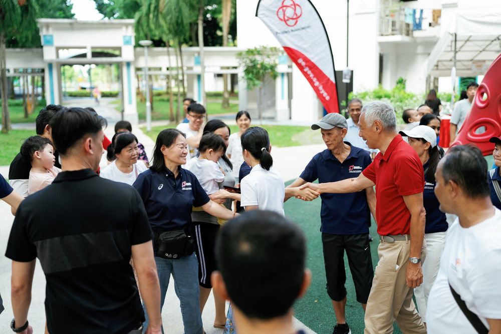 A group of people gathered outdoors for a training or team-building activity with red banners and foliage in the background.
