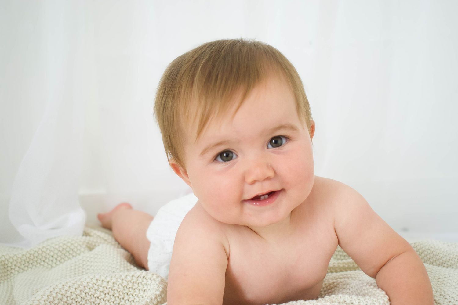 Baby in diaper smiles and plays on white blanket during photo session.