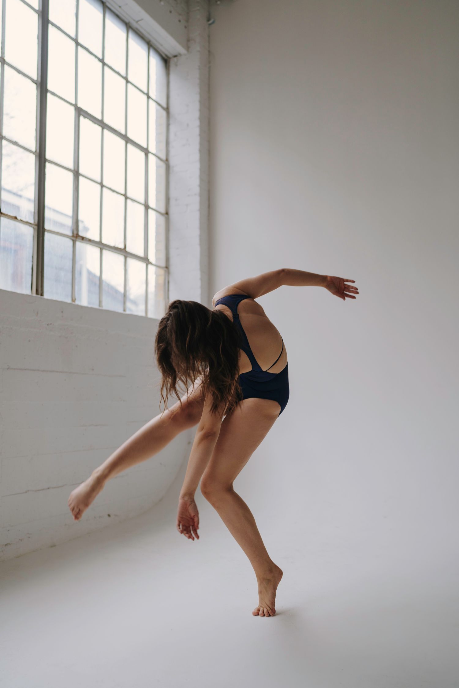 A figure in a dark outfit performs modern dance movements across a light-filled space with dramatic window shadows on the floor.