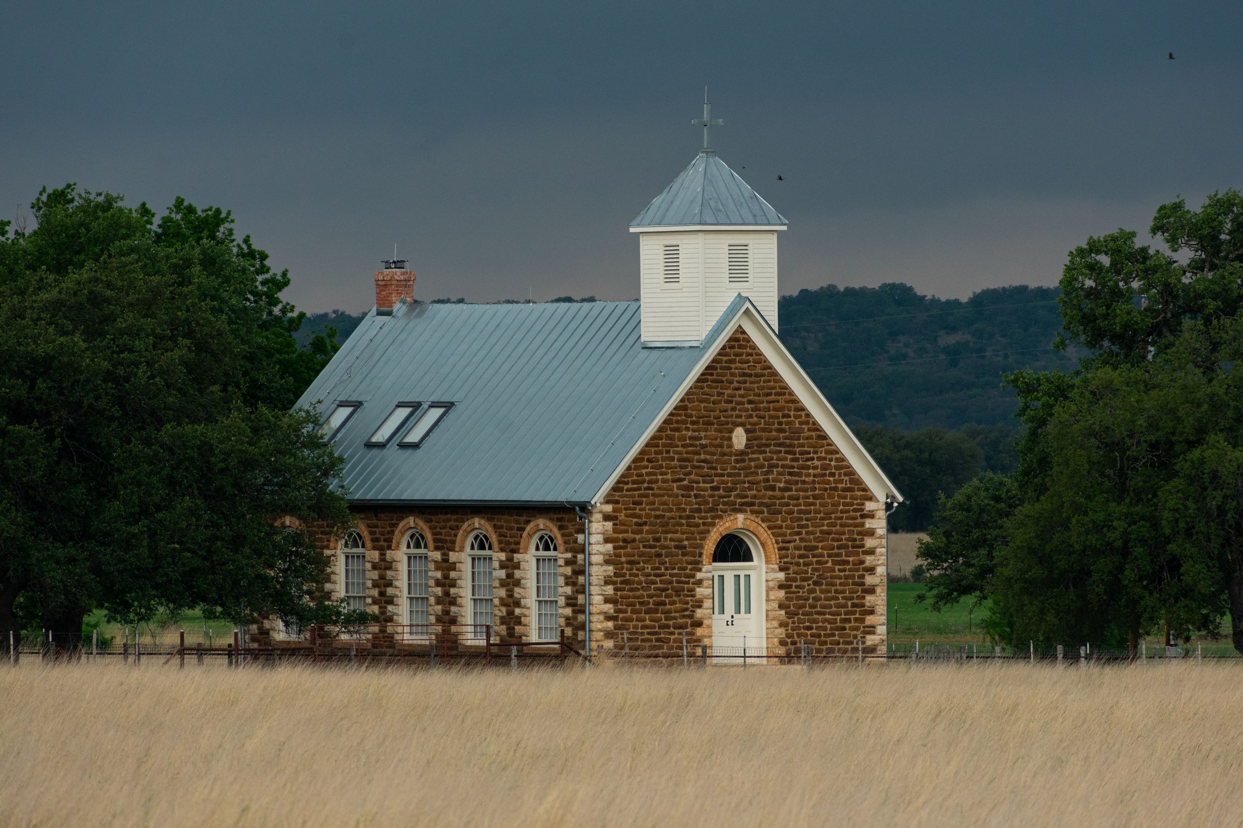 Rural church building with stone facade and white steeple set against dramatic sky.