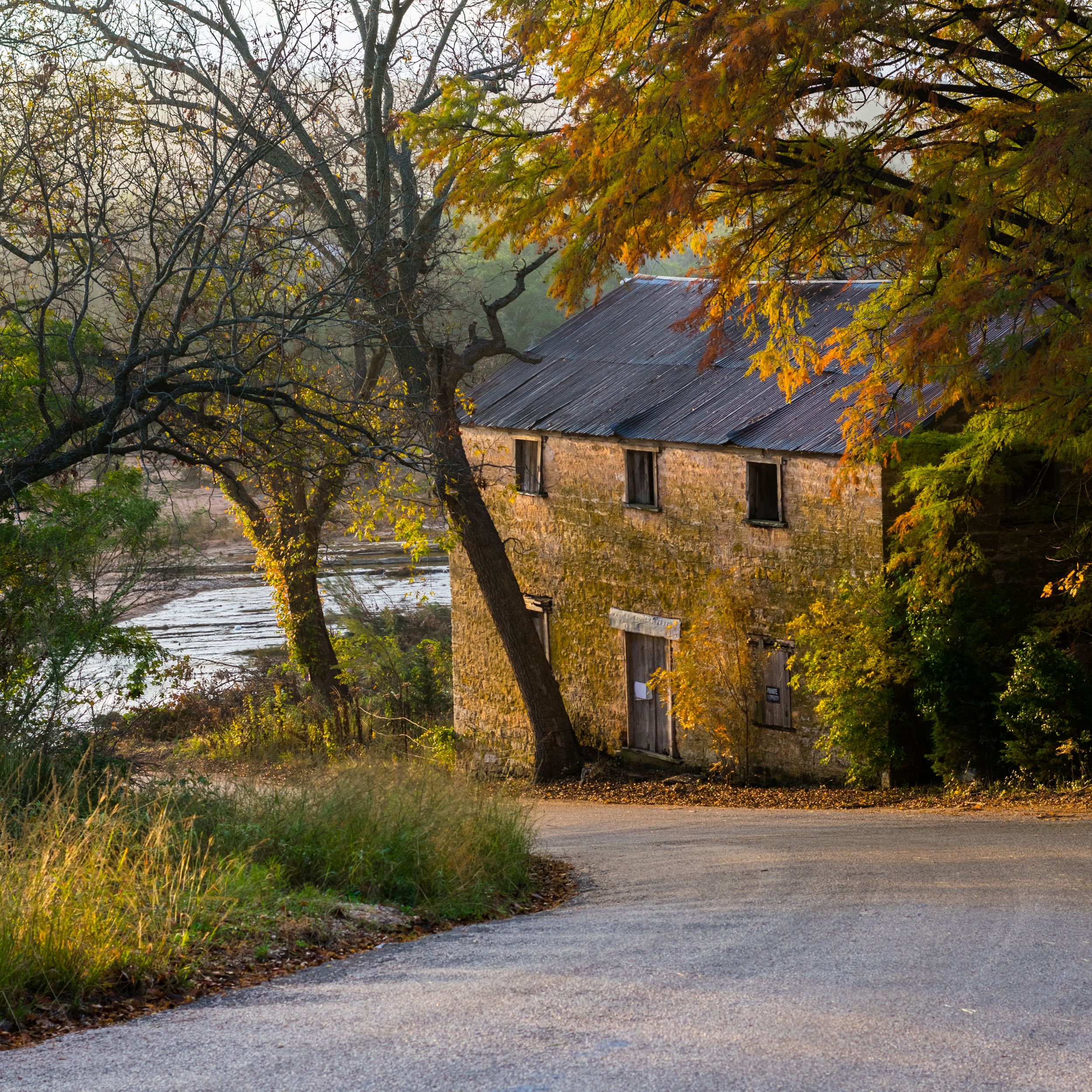 A winding gravel road leads to rustic stone buildings surrounded by autumn trees with golden foliage.