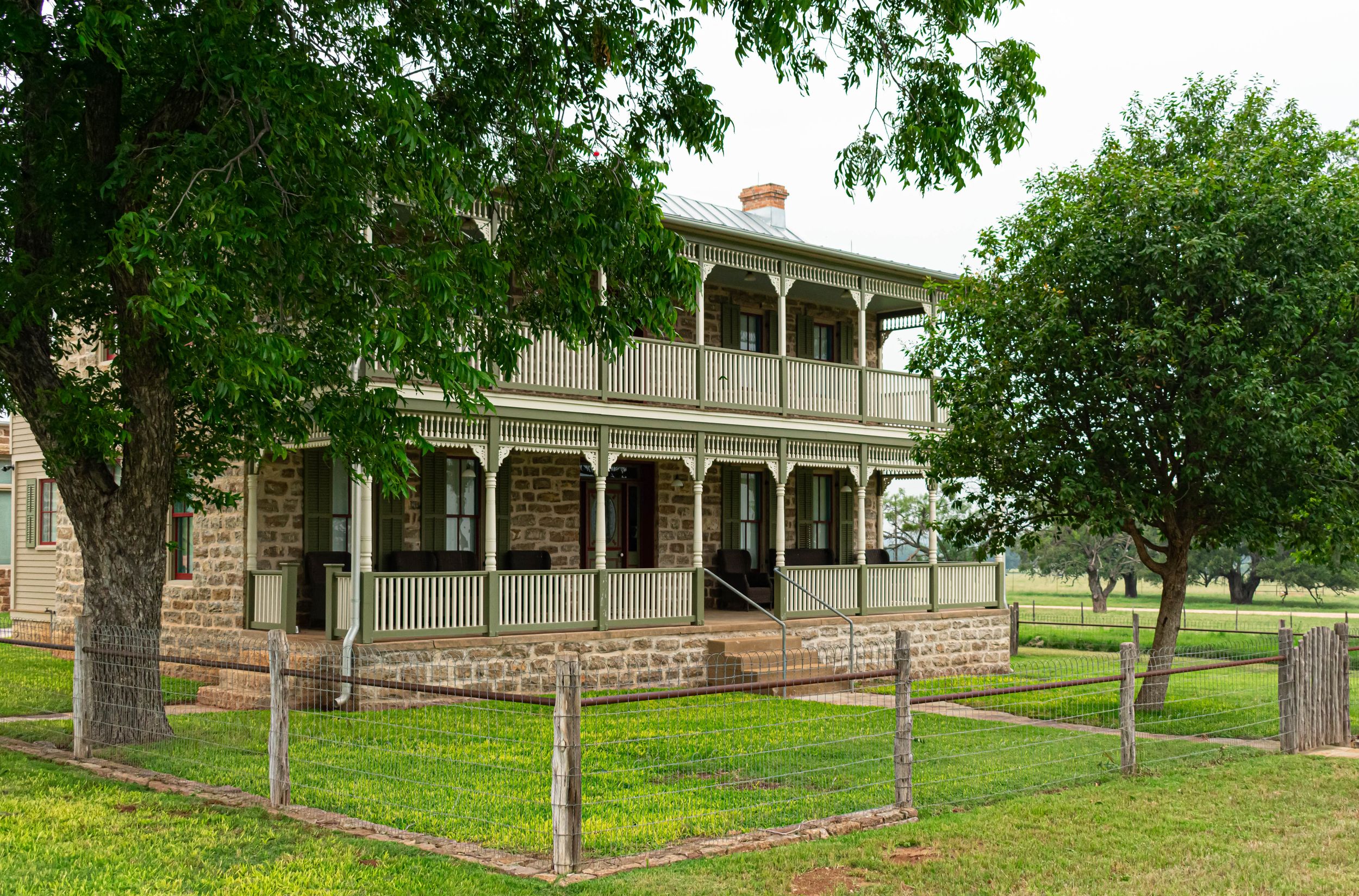 Historic stone building with wrap-around porch and mature trees on a manicured lawn.