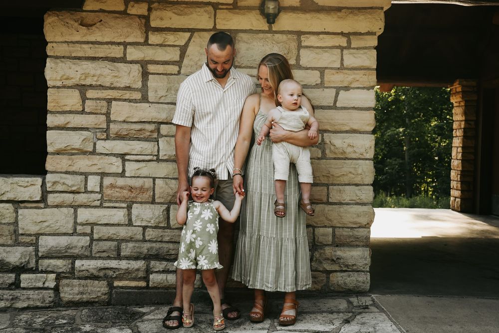 A family stands together against a stone wall background in coordinated neutral-toned outfits.