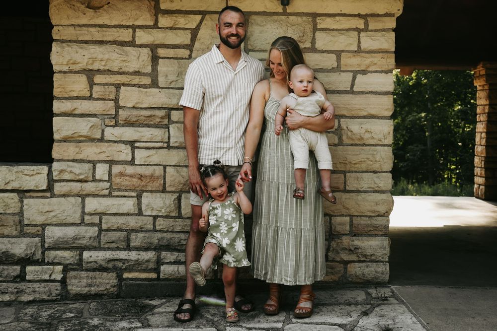 Family in neutral colored clothing stands against stone wall backdrop during outdoor photo session.