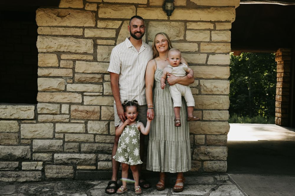 A family stands together against a stone wall backdrop while wearing casual summer attire.