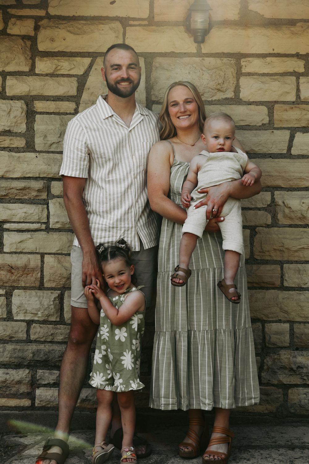 Family dressed in coordinating neutral tones poses against stone wall backdrop in casual outdoor photo session.