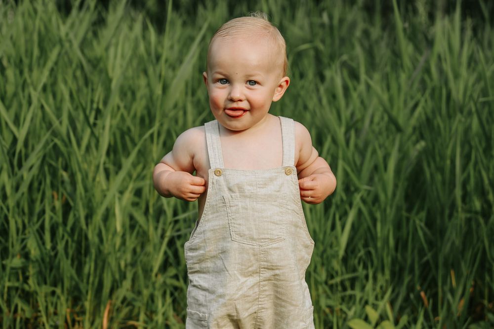 A small child wearing beige overalls stands in tall green grass on a sunny day while smiling.